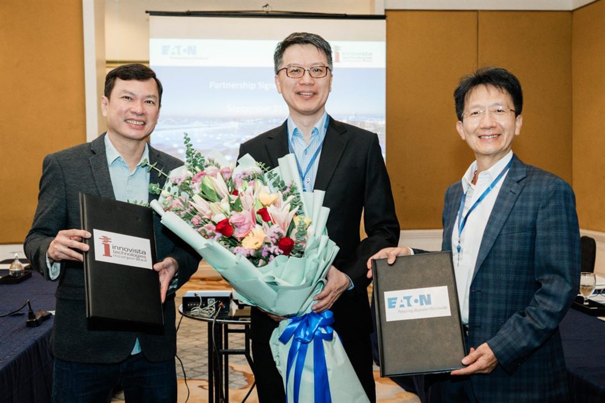  Three men in suits are posing for a photo. The man in the middle is holding a bouquet of flowers. The man on the left is holding a folder that says "Inovista". The man on the right is holding a folder that says "Eaton". The image is about Eaton 5A Advantage Series UPS in Indonesia.
